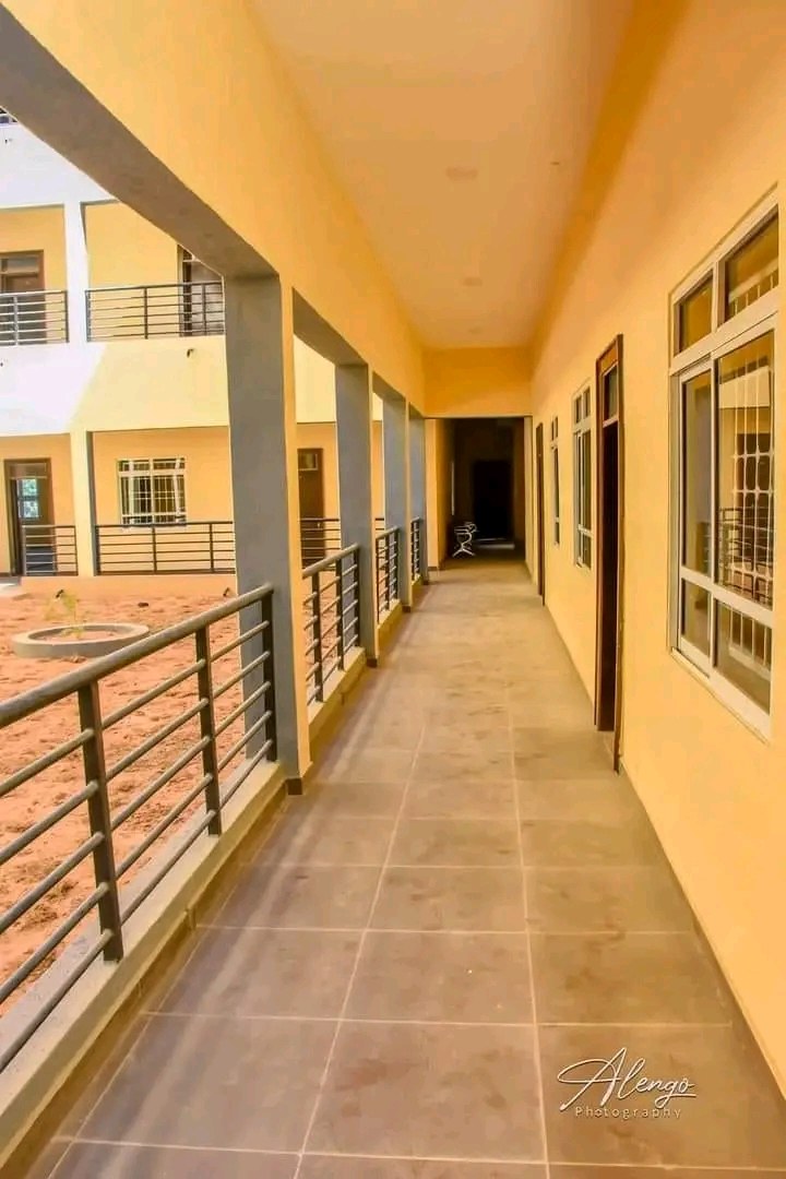 Ein mehrstöckiges Gebäude mit Fenstern, Türen, Säulen und Geländern, das einen langen gefliesten Flur und einen Balkon sowie andere Gebäude und einen klaren blauen Himmel zeigt, mit Text unten, der eine 3-Zimmer-Wohnung in Ntinda, Kampala zur Miete anbietet.