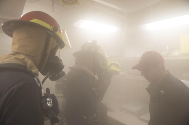 Feuerwehrleute in einem raucherfüllten Raum während einer Übung, einer hält eine Kamera, trägt Helme mit Lampen und Ausrüstung im Hintergrund sichtbar.
