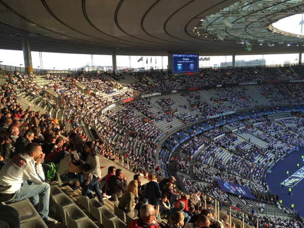 Eine große Menschenmenge in einem Stadion schaut einem Fußballspiel zu, mit einer Bühne, Fahnen, Stangen, einem Bildschirm und der Allianz Arena in München im Hintergrund.