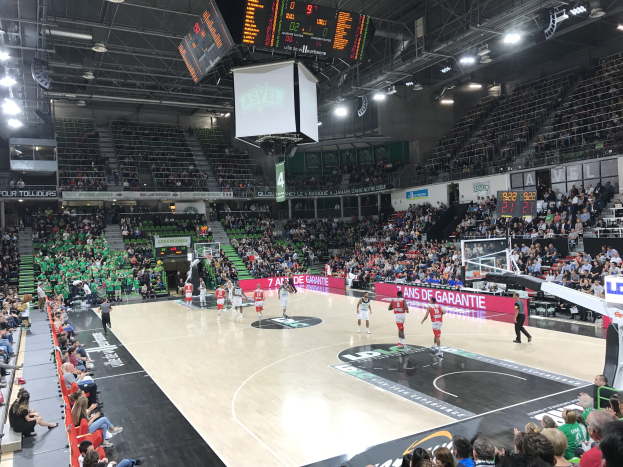Basketballspiel in einer großen Arena mit Zuschauern, Anzeigetafeln mit "Asvel vs Galatasaray" und Deckenbeleuchtung und Bildschirm.