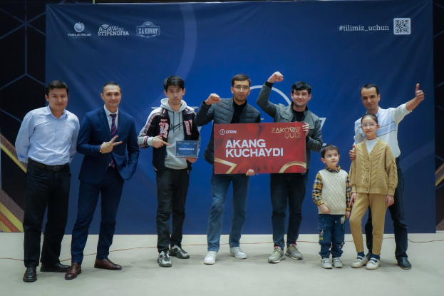 Eine Gruppe von Menschen auf einer Bühne, die eine Tafel mit der Aufschrift "Akang Kuchaydi - Zakovat Quiz" halten, mit einem Banner im Hintergrund.