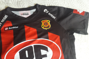 Ein schwarzes und rotes Fußballtrikot mit einer roten und schwarzen Streifen, einem Logo und Text, das auf einer weißen Oberfläche liegt.