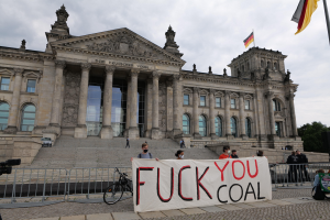 Gruppe von Menschen mit einem "Fuck You Coal"-Schild vor dem Reichstag in Berlin, mit architektonischen Details, einem Fahrrad, Bäumen, einer Fahnenstange und einem bewölkten Himmel.
