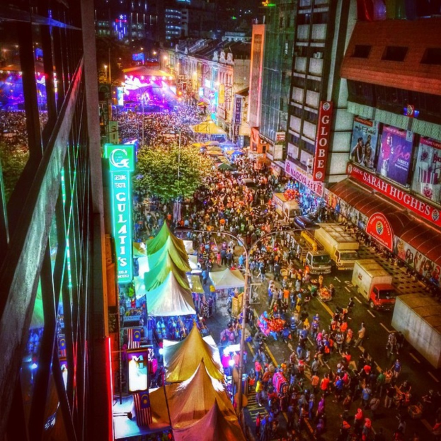 Ein volles Karnevalszelt mit Buden, Häusern, Gebäuden und Fahnen, mit beleuchteter Fläche im Hintergrund.