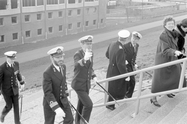 Schwarzes und weißes Foto einer Gruppe uniformierter Personen, die eine Treppe mit einem Geländer auf der rechten Seite hinuntergehen, vor einer Kulisse aus Gebäuden, Bäumen, Strommasten und einer Straße.