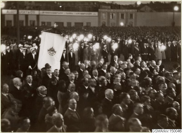 Schwarzes und weißes Foto einer großen Menge vor einem Gebäude, mit einer Person, die eine Fahne hält, Polen und Lichter im Hintergrund und Text unten mit der Aufschrift "Gustav Klimts Rede an der Berliner Mauer".