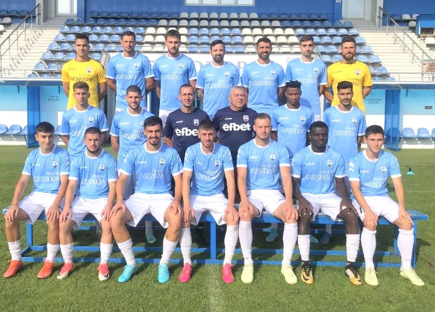 Gruppe von Männern auf einem Fußballfeld, einige sitzen auf Bänken und einige stehen auf dem Rasen, mit Stühlen, Treppen, Geländern, Wänden und Fenstern im Hintergrund.