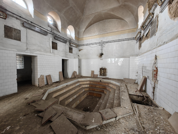 Ein verlassenes Gebäude mit einem großen runden Pool in der Mitte, gefliesten Wänden, Fenstern auf beiden Seiten und hölzernen Gegenständen, die den Pool umgeben, mit einem Wasserzeichen auf dem Bild.