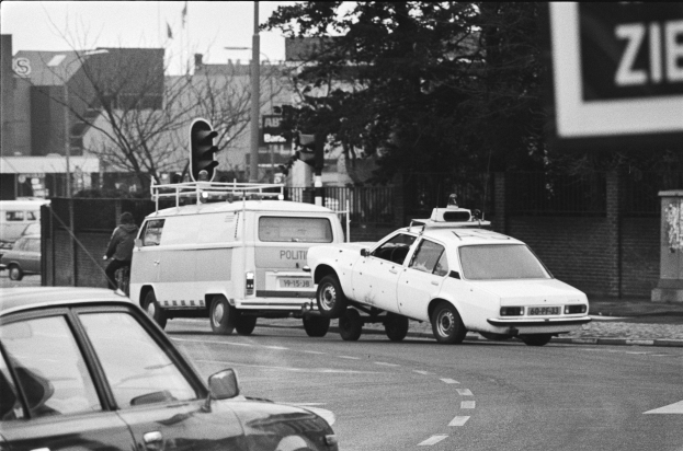 Schwarzes und weißes Foto eines Autos, das von einem Polizeiwagen auf einer Stadtstraße mit anderen Fahrzeugen, einem Fußgänger, Ampeln, Pfählen, Bäumen, Gebäuden und einem Zaun im Hintergrund geschleppt wird.