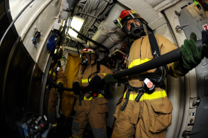 Eine Gruppe von Feuerwehrleuten in Helmen und Handschuhen hält einen Schlauch im Heck eines Flugzeugs, mit Kabeln, Lampen und anderen Gegenständen im Hintergrund und Text am unteren Bildrand.