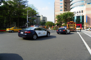 Ein Polizeiwagen fährt eine Straße mit hohen Gebäuden, Bäumen, Laternen, Verkehrsampeln und Menschen unter einem bewölkten Himmel.