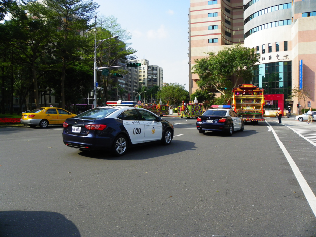 Ein Polizeiwagen fährt eine Straße mit hohen Gebäuden, Bäumen, Laternen, Verkehrsampeln und Menschen unter einem bewölkten Himmel.