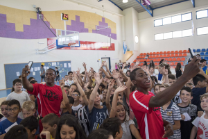 Kinder auf einem Basketballplatz mit Handys stehend, mit einem Tor, einem Basketballkorb, einer Uhr, Papieren auf einer Tafel, Deckenlampen, Stühlen und Fenstern im Hintergrund.