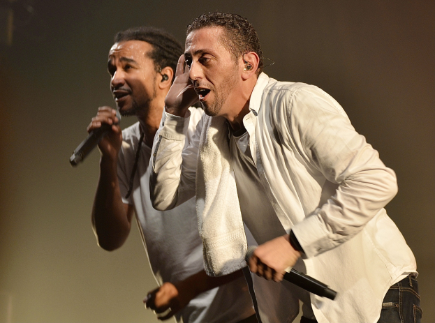 Zwei Männer in legerer Kleidung stehen nebeneinander auf einer Bühne, jeder hält ein Mikrofon in der Hand.