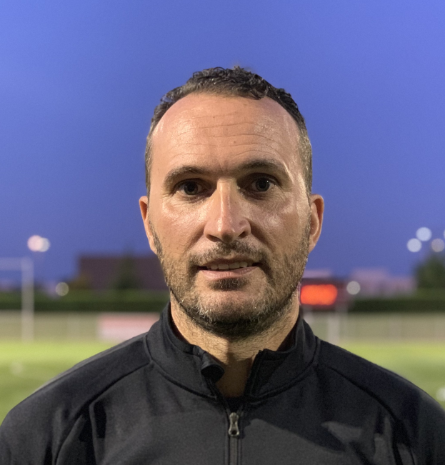 Ein Mann in einer schwarzen Jacke mit Logo steht auf einem Fussballfeld und scheint ein Trainer zu sein, mit einem unscharfen Hintergrund.