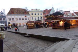 Ein lebendiger Weihnachtsmarkt in Tallinn, Estland, mit Menschen um beleuchtete Stände, festlicher Dekoration, Gebäuden mit Fenstern im Hintergrund, Treppen, die zum Markt führen, und Töpfen mit Pflanzen.
