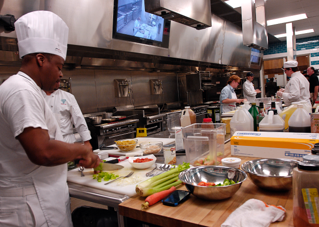 Eine Gruppe von Köchen in einer Küche beim Zubereiten von Essen auf einem Schneidbrett, wobei ein Koch im Vordergrund in einer weißen Schürze und einer Mütze steht und ein Messer hält und von verschiedenen Utensilien, Schüsseln, Gemüse, einem Mobiltelefon, Flaschen und anderen Gegenständen umgeben ist, mit einem Fernseher und Deckenleuchten im Hintergrund.