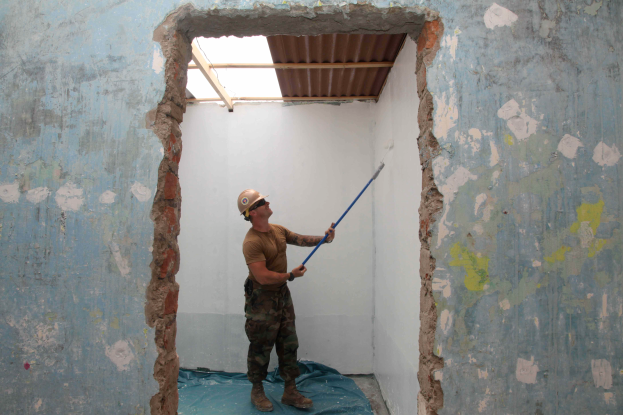 Ein Mann in einem Helm hält einen Stock, steht in einem Raum mit einem Loch in der Wand und einem Dach darüber, umgeben von Putzwerkzeugen und -materialien.
