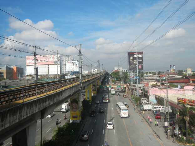 Eine belebte Stadtstraße mit Fahrzeugen, einer Überführung, Strommasten, Bäumen, Gebäuden, Werbetafeln und einem bewölkten Himmel.