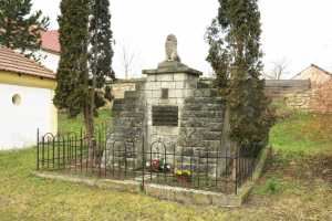 Ein steinernes Denkmal auf einer grünen Wiese, umgeben von einem Metallzaun und geschmückt mit Blumensträußen, mit Bäumen, Häusern und einer Wand im Hintergrund unter einem klaren blauen Himmel dient als Holocaust-Mahnmal in Sibiu, Rumänien.