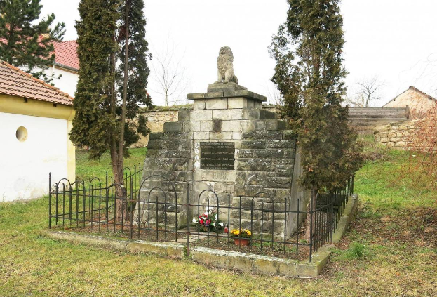 Ein steinernes Denkmal auf einer grünen Wiese, umgeben von einem Metallzaun und geschmückt mit Blumensträußen, mit Bäumen, Häusern und einer Wand im Hintergrund unter einem klaren blauen Himmel dient als Holocaust-Mahnmal in Sibiu, Rumänien.