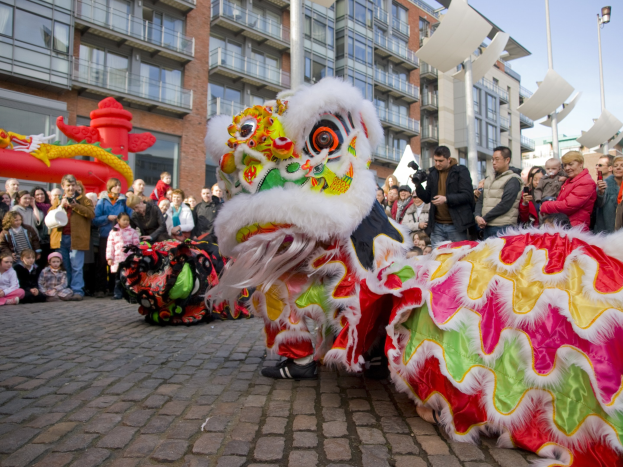 Ein lebendiges chinesisches Neujahrsfest in Amsterdam mit einem Löwen tanzen vor einer Zuschauermenge, einige halten Kameras, vor einer Kulisse aus Gebäuden, Laternenmasten und einem klaren blauen Himmel.