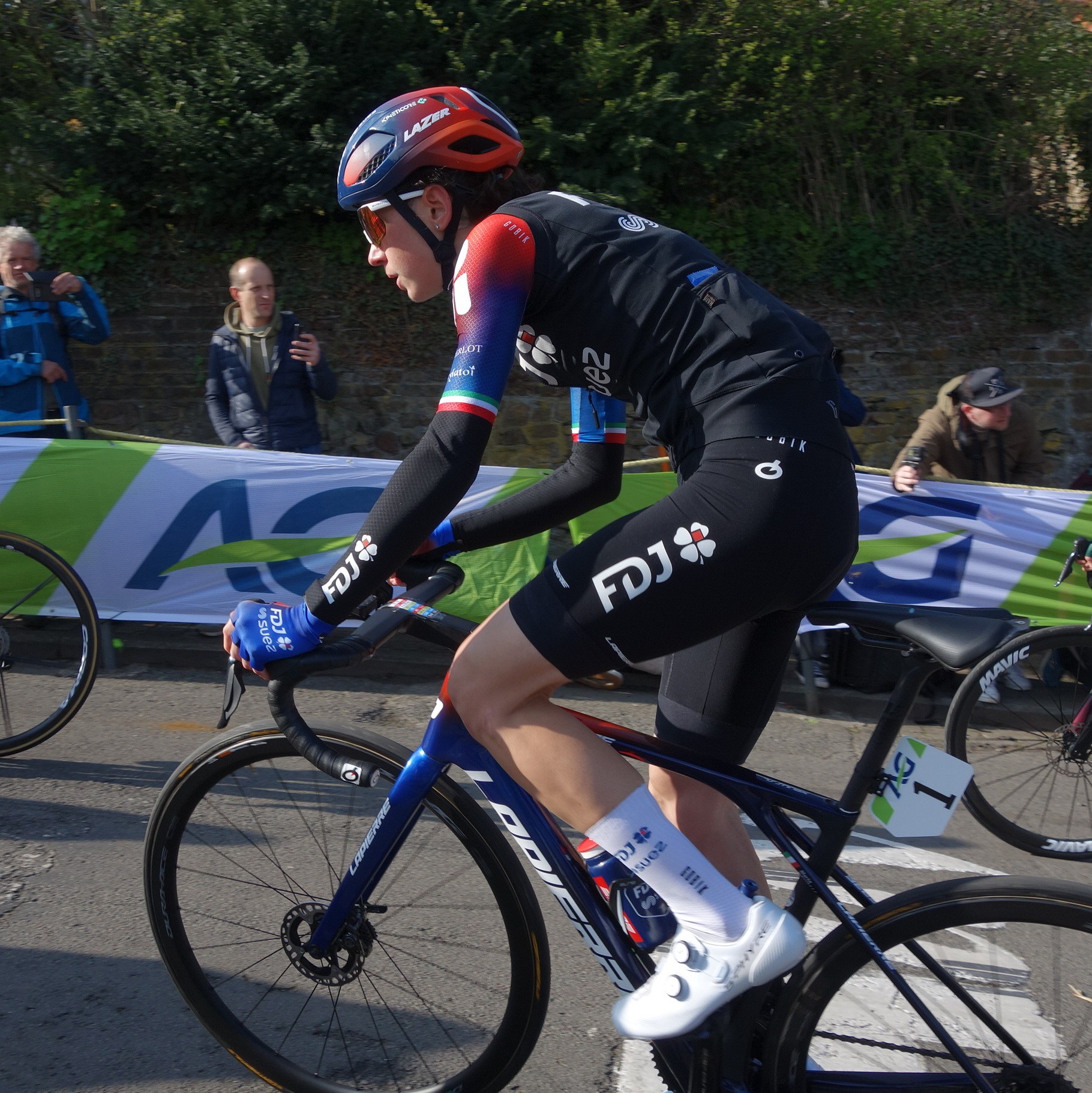Professionelle Radrennfahrerin Katie Archibald trägt einen Helm, eine Schutzbrille, Handschuhe und Schuhe und fährt ein Fahrrad auf einer Straße an einer Zuschauermenge vorbei, mit einem Banner, einer Wand, Bäumen und einem Gebäude im Hintergrund.