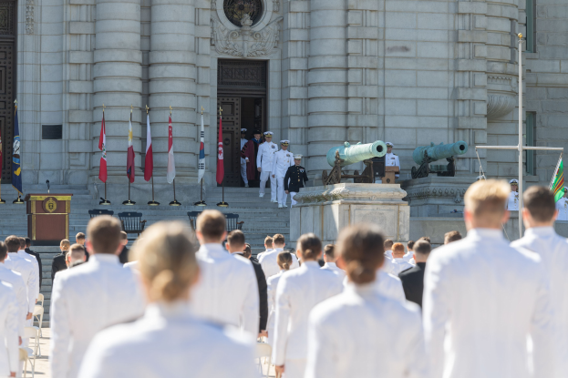 Gruppe von Menschen in weißen Marineuniformen, die auf einer Treppe vor einem Gebäude mit Säulen, einer Tür und Fahnen während einer Abschlussfeier stehen.