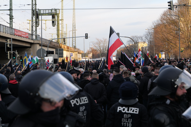Eine große Gruppe von Polizisten steht vor einer Menge, die Fahnen schwingt und Helme trägt, mit Fahrzeugen, Infrastruktur und Gebäuden im Hintergrund.