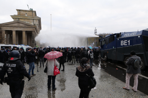 Eine Gruppe von Menschen vor einer Reihe von Polizeibeamten in Berlin, einige mit Helmen und Schirmen, mit Fahrzeugen, einem Gebäude mit Säulen, Bäumen und Laternen im Hintergrund.