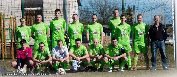 Eine Gruppe von Männern in grünen T-Shirts steht auf einem Fußballfeld, einige sind in der Hocke, mit einem Ball und einem Torpfosten; der Hintergrund zeigt Bäume, Gebäude und Fahrzeuge.
