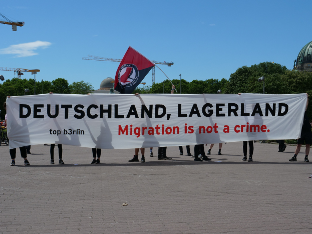 Gruppe von Menschen mit einem Transparent mit der Aufschrift 'Deutschland, Lagerland Migration ist kein Verbrechen' und einer Flagge, im Hintergrund sind Straßenlaternen, Bäume, Kräne, ein Gebäude und ein bewölkter Himmel zu sehen.