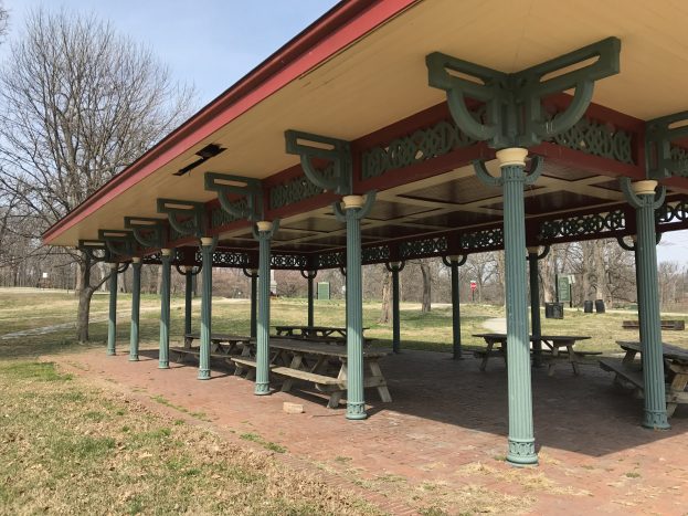 Ein hölzerner Pavillon mit Picknicktischen steht auf einer grünen Wiese umgeben von Bäumen unter einem klaren blauen Himmel.