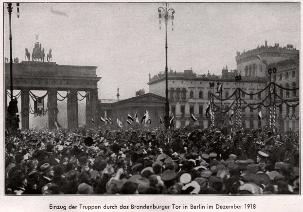 Menge von Menschen mit Hüten und einigen, die Fahnen halten, vor dem Brandenburger Tor in Berlin, Deutschland, 1918, mit dem Torbogen, den Statuen und den umliegenden Gebäuden im Hintergrund.