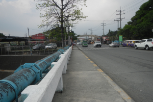 Stadtstraße mit fahrenden Autos, eine Brücke im Hintergrund, Strommasten mit Drähten, Bäume, Gebäude und ein bewölkter Himmel, mit einem blauen Rohr an der Straßenrand.