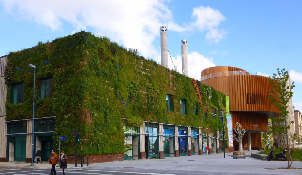 Ein großes Gebäude mit einer grünen Wand an der Seite, umgeben von Straßenmasten, Straßenlaternen, Verkehrspfosten, Verkehrsampeln, Kraftfahrzeugen, Fußgängern, Bäumen und einer bewölkten Himmel, als Teil des Green Building Project.