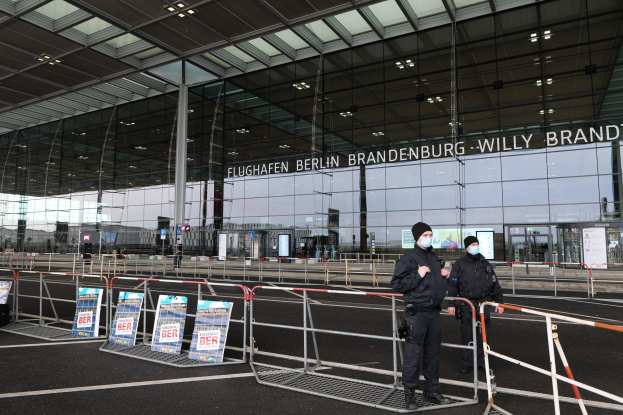 Zwei Polizisten in schwarzer Uniform stehen vor einem Glasgebäude mit Text, mit Absperrungen und Schildern im Vordergrund.