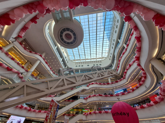Ein Einkaufszentrum mit pinken und weißen Ballons, Rolltreppen, Geländern, Schildern, Lichtern und einer Glasdecke geschmückt zum Valentinstag.