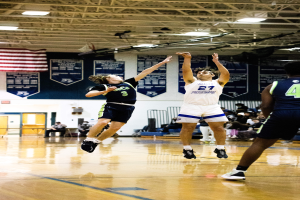 Eine Gruppe von Menschen, die Basketball in einer Turnhalle spielen, mit Zuschauern im Hintergrund, Bannern und einer Flagge an der Wand und einer Decke mit Stangen und Lampen darüber.