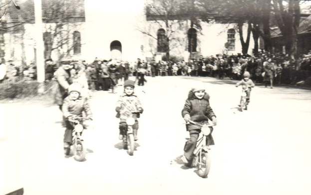 Schwarze-Weiß-Foto von Kindern, die auf der Straße Fahrräder fahren, mit einem Gebäude und Bäumen im Hintergrund.