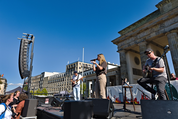 Gruppe von Menschen, die auf einer Bühne Musik spielen mit dem Brandenburger Tor und Berliner Gebäuden im Hintergrund.