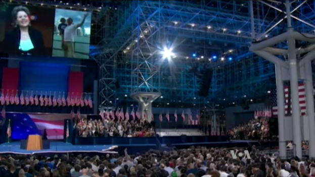 Große Menschenmenge vor einer Bühne mit Podium, Mikrofon, Flaggen, einem Bildschirm und Beleuchtungsausrüstung, was auf eine Präsidentschaftsdebatte hindeutet.