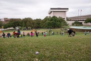 Gruppe von Menschen beim Ostereier-Spiel auf einer grünen Wiese mit Bäumen, Gebäuden, Fahnen, einem Brunnen und einem bewölkten Himmel im Hintergrund.