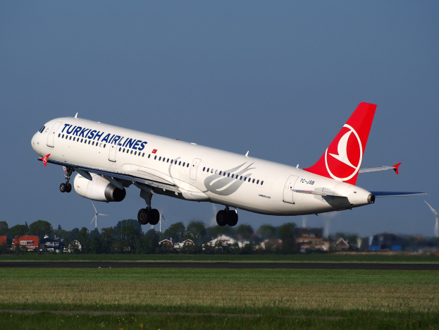 Turkish Airlines Airbus A330-300 steigt von der Startbahn des Frankfurter Flughafens auf, mit Gras darunter, Gebäuden und Bäumen im Hintergrund, fernen Windmühlen und einem klaren blauen Himmel.
