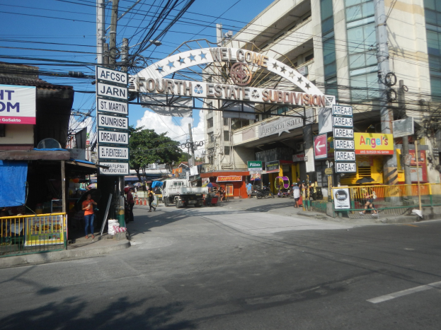 Eine belebte Stadtstraße mit fahrenden Fahrzeugen, Fußgängern auf dem Gehweg, einem Torbogen mit Text, Gebäuden, elektrischen Masten mit Drähten, Bäumen und einem bewölkten Himmel, mit einem "Fourth Estate Subdivision"-Straßenschild in der Mitte.