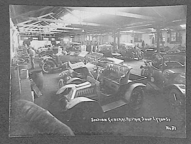 Schwarzes und weißes Foto einer frühen 20. Jahrhundert-Autowerkstatt in Lyon, Frankreich, das mehrere Fahrzeuge, Menschen, verstreute Werkzeuge und eine sichtbare Decke mit Text am Boden zeigt.