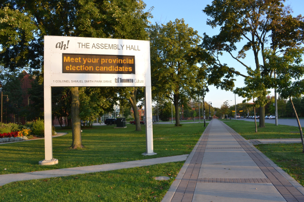 Tafel auf Rasen neben einem Weg mit dem Text "The Assembly Hall - Treffen Sie Ihre Provincial Election Candidates" umgeben von Bäumen, Blumen, Straßenlaternen, Fahrzeugen, einem Gebäude und einem bewölkten Himmel.