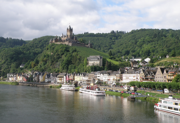 Ein malerischer Blick auf den Rhein in Deutschland mit einer Burg auf einem Hügel, Booten auf dem Fluss und Fahrzeugen auf der Straße sowie einem bewölktem Himmel.