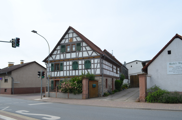 Ein altes Rathauses mit weißer und schwarzer Fassade an einer Straßenkreuzung, umgeben von Pflanzen mit Blumen, einem Schild, einem Straßenschild, einem Verkehrssignal und einer Tafel mit Text, unter einem bewölkten Himmel.