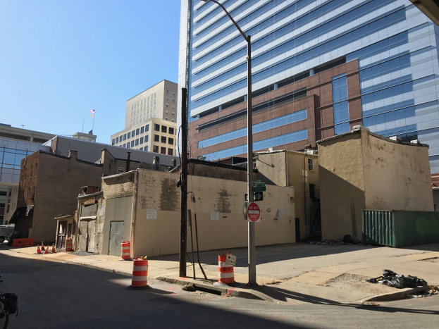 Eine Stadtstraße mit einem geparkten Fahrrad, umgeben von Gebäuden, Straßenmöbeln, Kraftfahrzeugen und einem demolierten Gebäude im Hintergrund unter einem klaren blauen Himmel.
